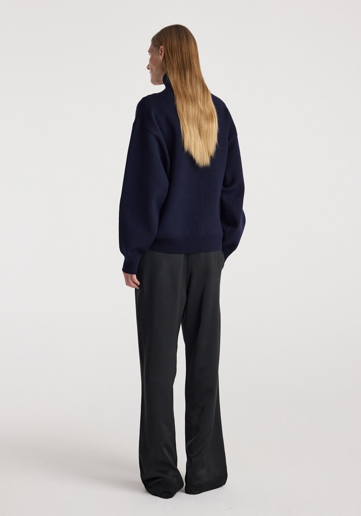 Woman shown from behind wearing a navy sculptural compact turtleneck with long sleeves paired with black wide-leg pants against a plain light background.