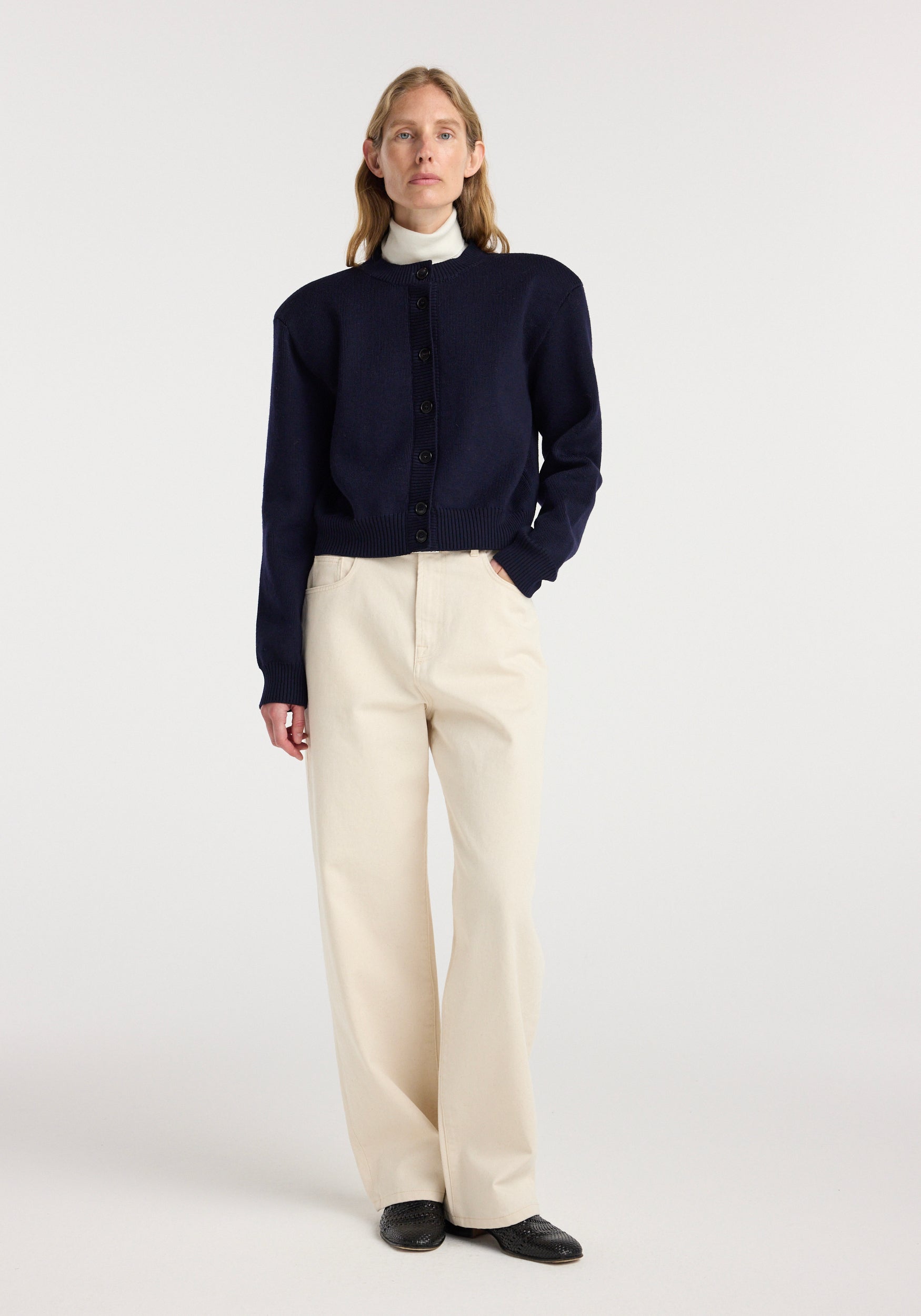 Woman wearing a navy Statement shoulder cardigan | navy with button front, styled with a white turtleneck and beige wide-leg pants against a plain background.