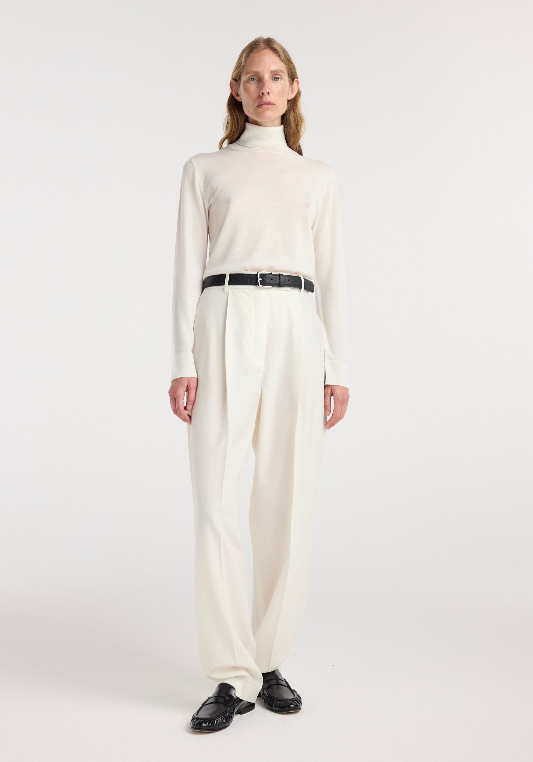 Model wearing a fine-knit wool cashmere turtleneck in off white paired with high-waisted cream trousers, a black belt, and black loafers, standing against a plain background.