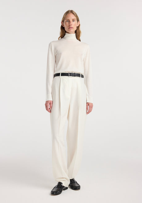 Model wearing a fine-knit wool cashmere turtleneck in off white paired with high-waisted cream trousers, a black belt, and black loafers, standing against a plain background.