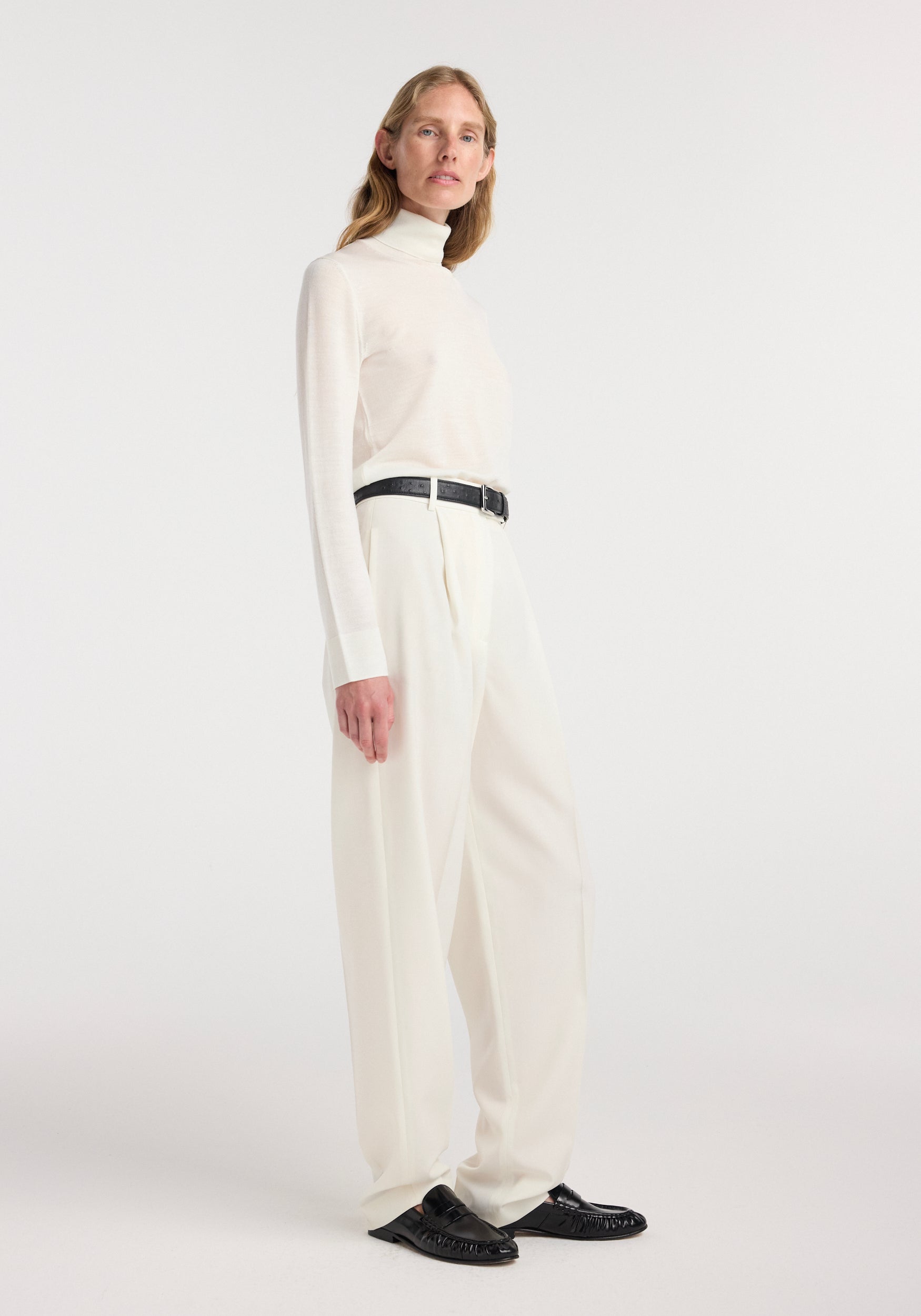 Woman wearing a fine-knit wool cashmere turtleneck | off white paired with high-waisted off-white trousers and black belt, standing against a plain white background.