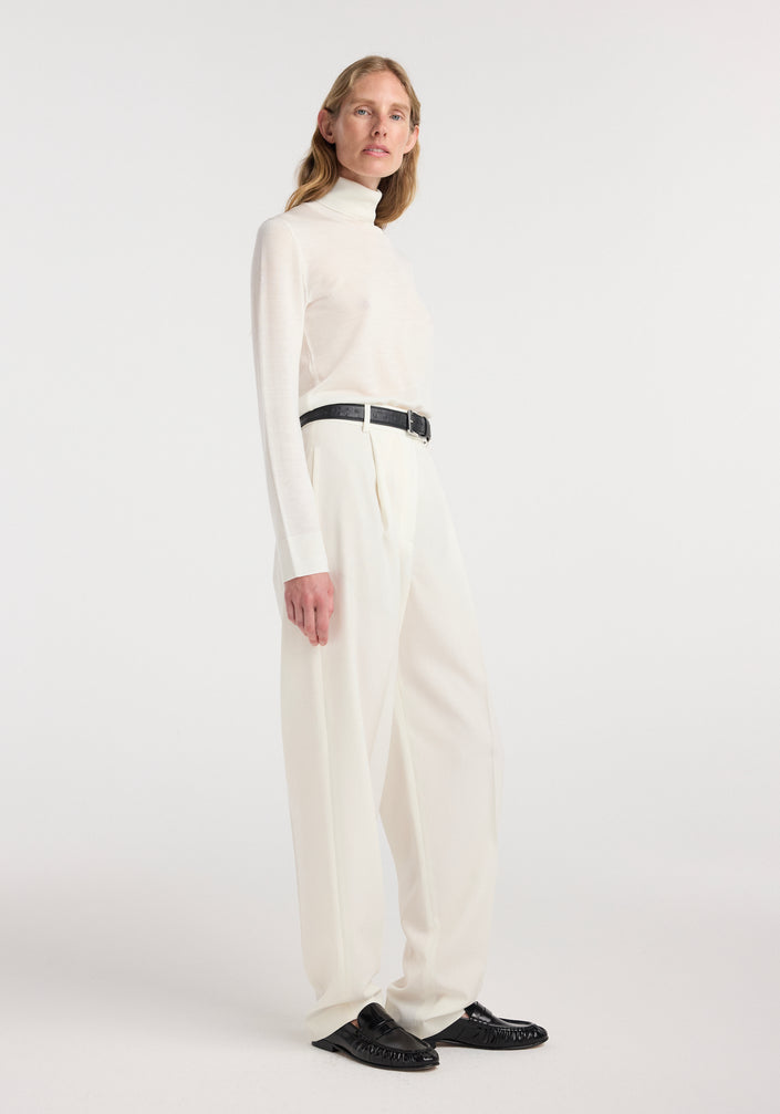 Model wearing a fine-knit wool cashmere turtleneck in off white paired with high-waisted cream trousers, a black belt, and black loafers, standing against a plain background.