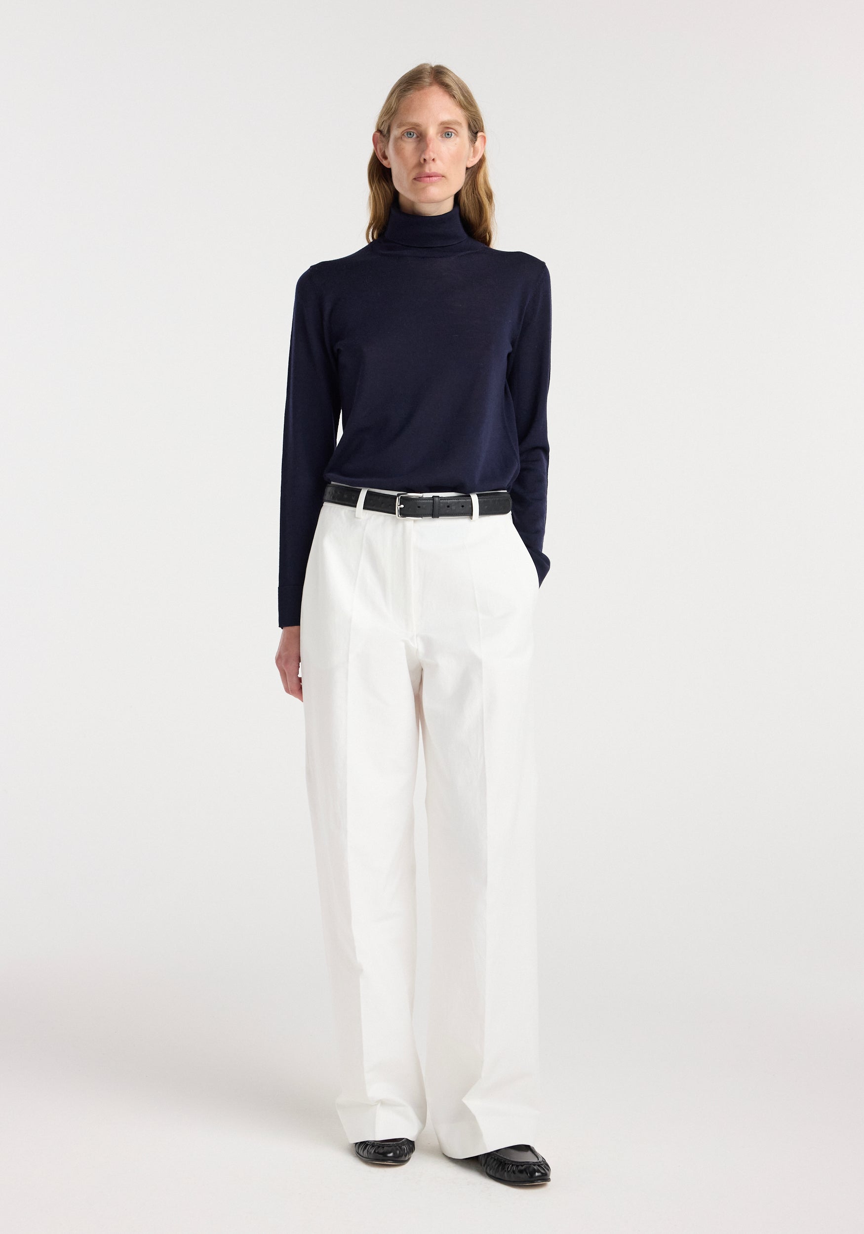 Model wearing a fine-knit wool cashmere turtleneck in navy paired with high-waisted white trousers, black belt, and black shoes, standing against a plain white background.