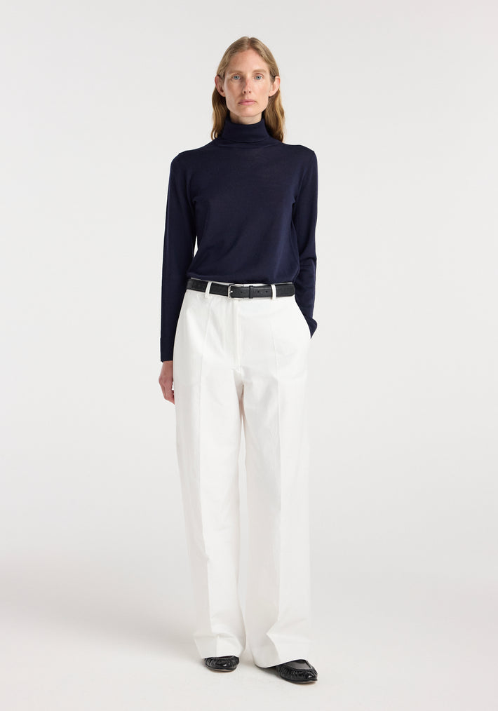Model wearing a fine-knit wool cashmere turtleneck in navy paired with high-waisted white trousers, black belt, and black shoes, standing against a plain white background.