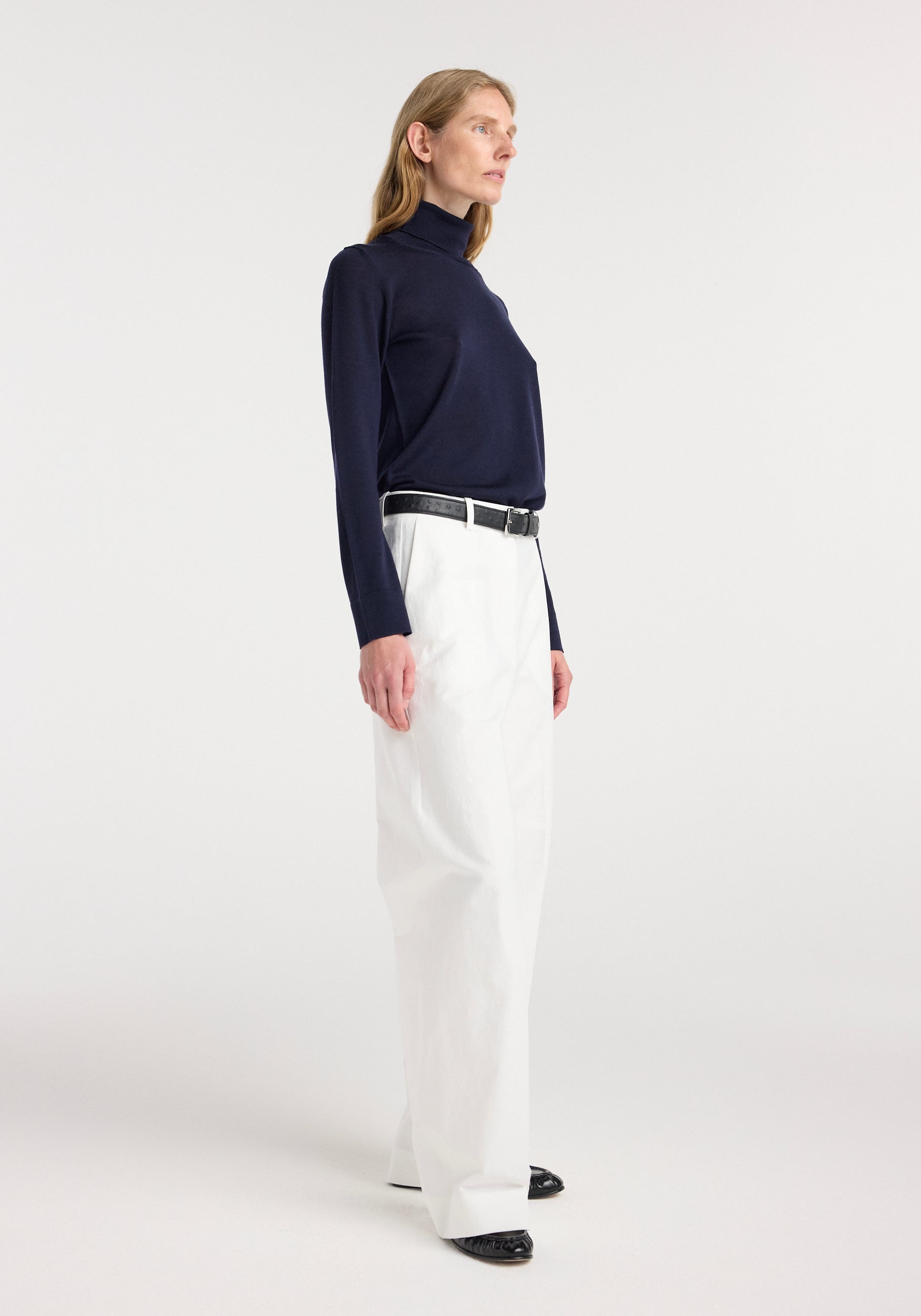 Woman wearing a navy fine-knit wool cashmere turtleneck with long sleeves, paired with high-waisted white trousers and a black belt, standing against a plain background.