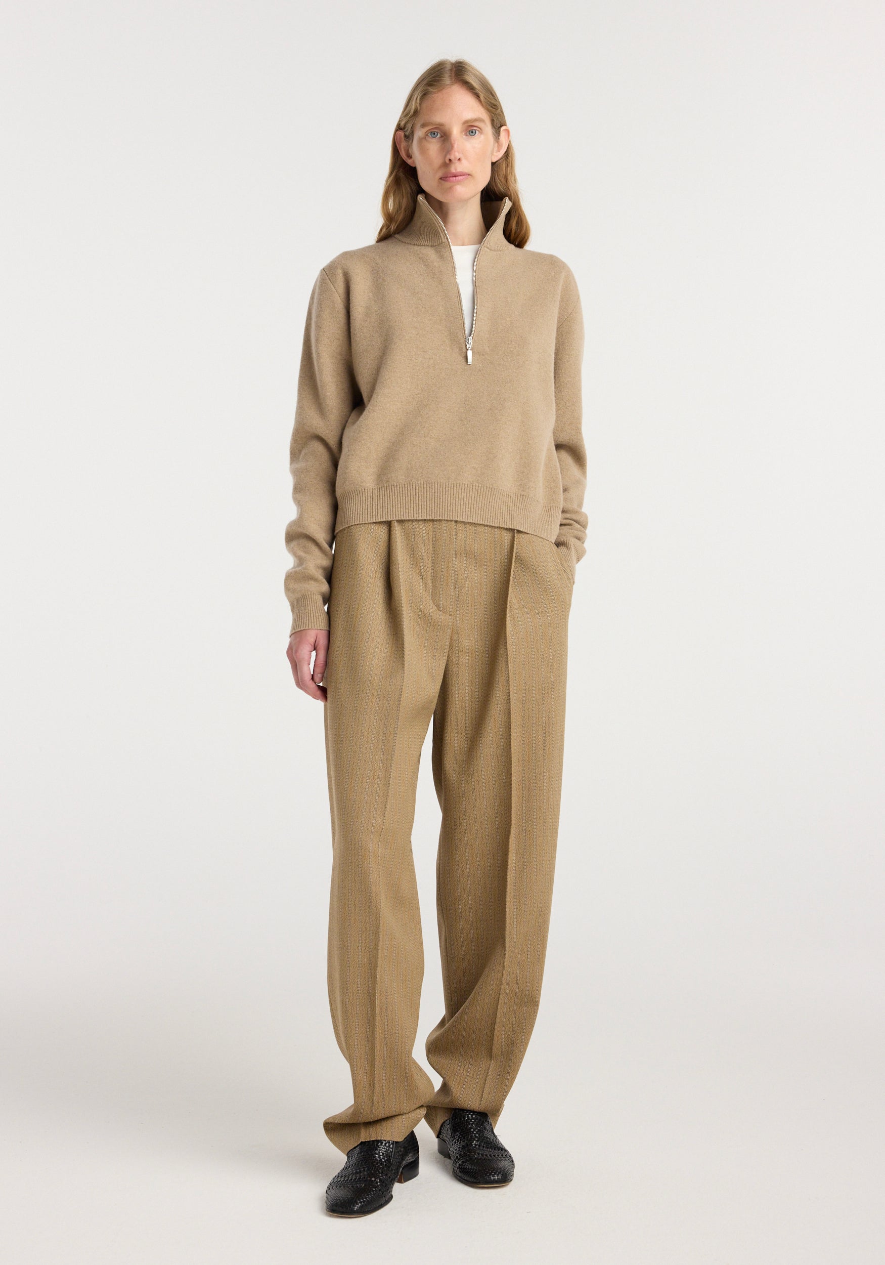 A model wearing beige single pleat tapered trousers with a subtle heritage stripe pattern paired with a light brown zip-up sweater and black shoes, showcasing Daria - Single pleat tapered trousers | heritage stripe.