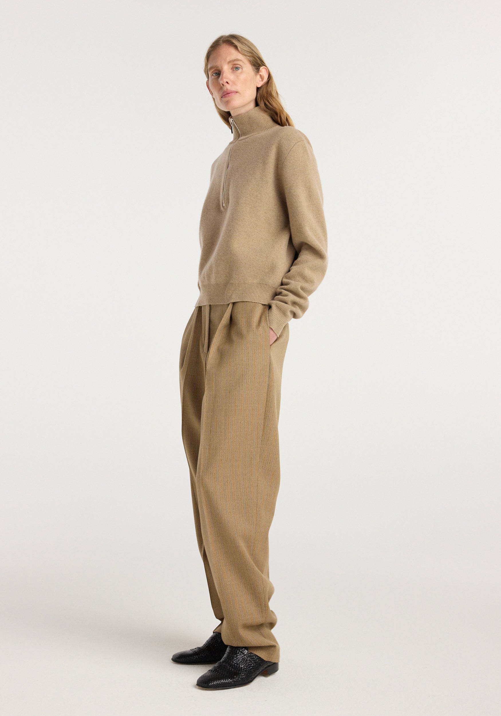 Woman wearing beige high-neck sweater paired with Daria - Single pleat tapered trousers | heritage stripe in a light brown tone, styled with black textured shoes against a plain background.
