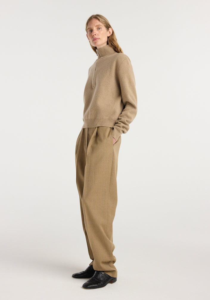 A model wearing beige single pleat tapered trousers with a subtle heritage stripe pattern paired with a light brown zip-up sweater and black shoes, showcasing Daria - Single pleat tapered trousers | heritage stripe.