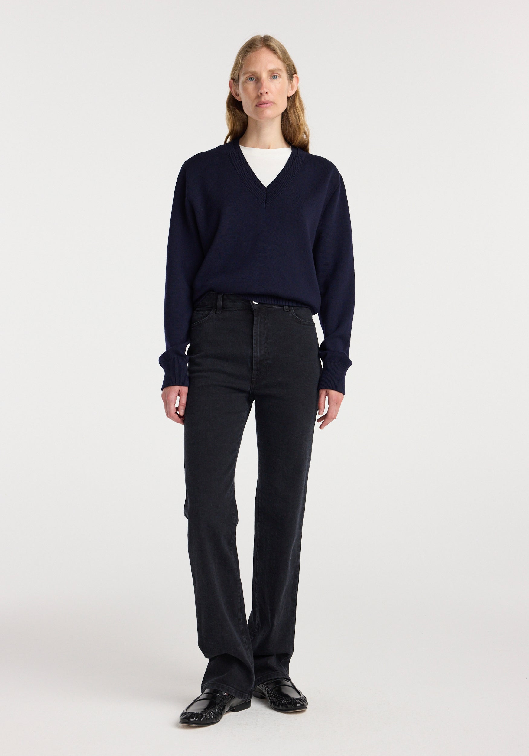 Woman wearing Lino - High-rise straight denim | black paired with a navy blue v-neck sweater and black shoes, standing against a plain white background.