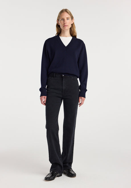 Woman wearing Lino - High-rise straight denim | black paired with a navy blue v-neck sweater and black shoes, standing against a plain white background.