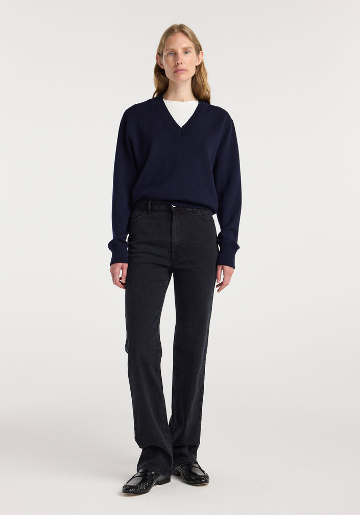 Woman wearing a navy double v-neck jumper with long sleeves over a white top, paired with black high-waisted jeans and black shoes, standing against a plain white background.