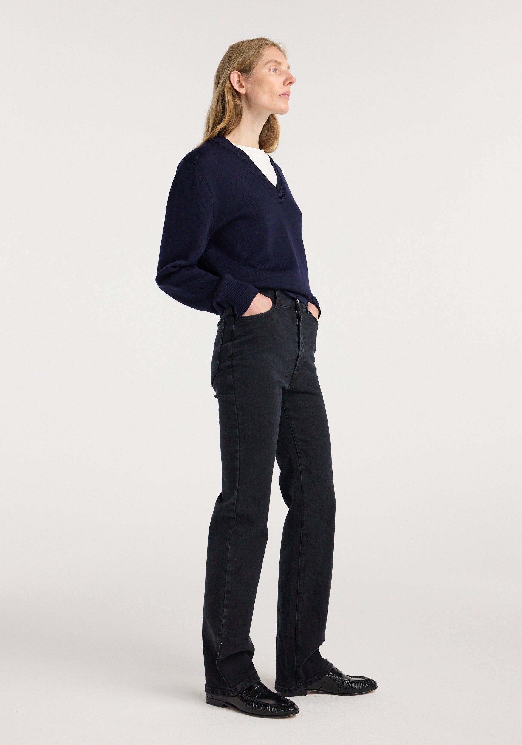 Woman wearing Lino - High-rise straight denim | black jeans paired with a navy sweater and black shoes standing against a plain light background with hands in pockets.