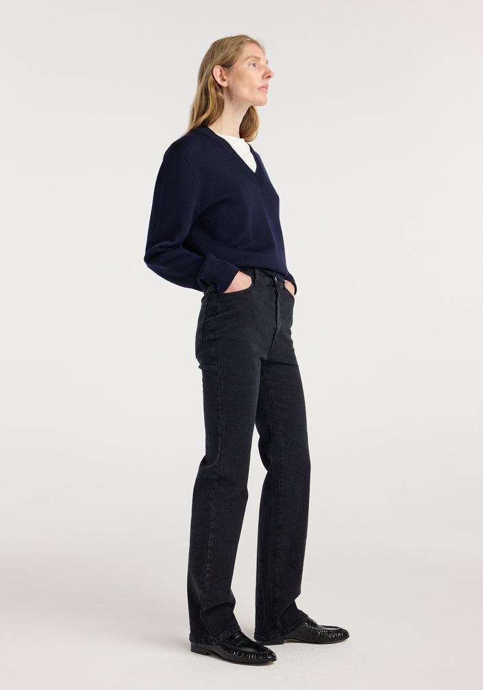 Woman wearing Lino - High-rise straight denim | black paired with a navy blue v-neck sweater and black shoes, standing against a plain white background.