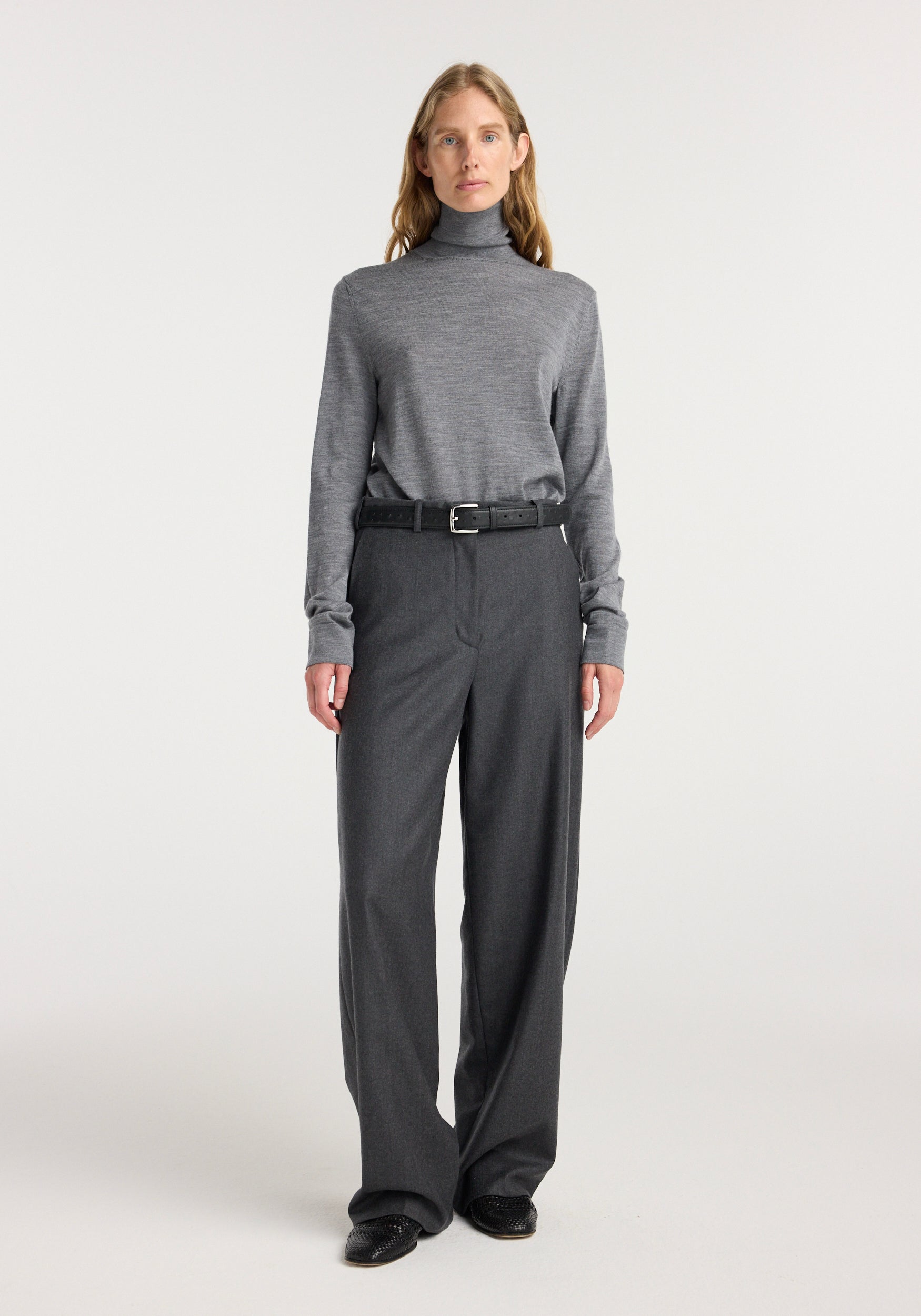 Woman wearing a grey turtleneck sweater paired with wide leg trousers | dark grey melange and a black belt, standing against a plain light background.