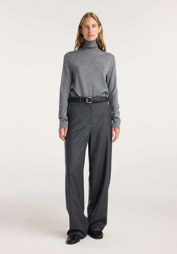 Woman wearing a grey turtleneck sweater paired with wide leg trousers | dark grey melange and a black belt, standing against a plain light background.
