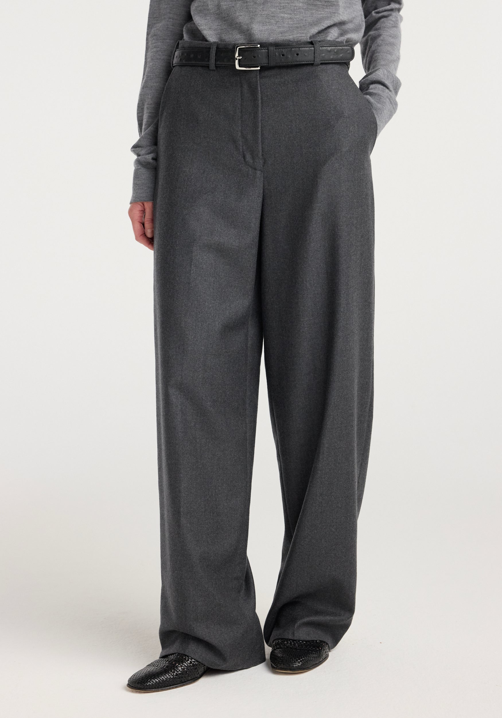 Front view of a person wearing wide leg trousers | dark grey melange styled with a matching grey top, black belt, and black shoes against a plain background.