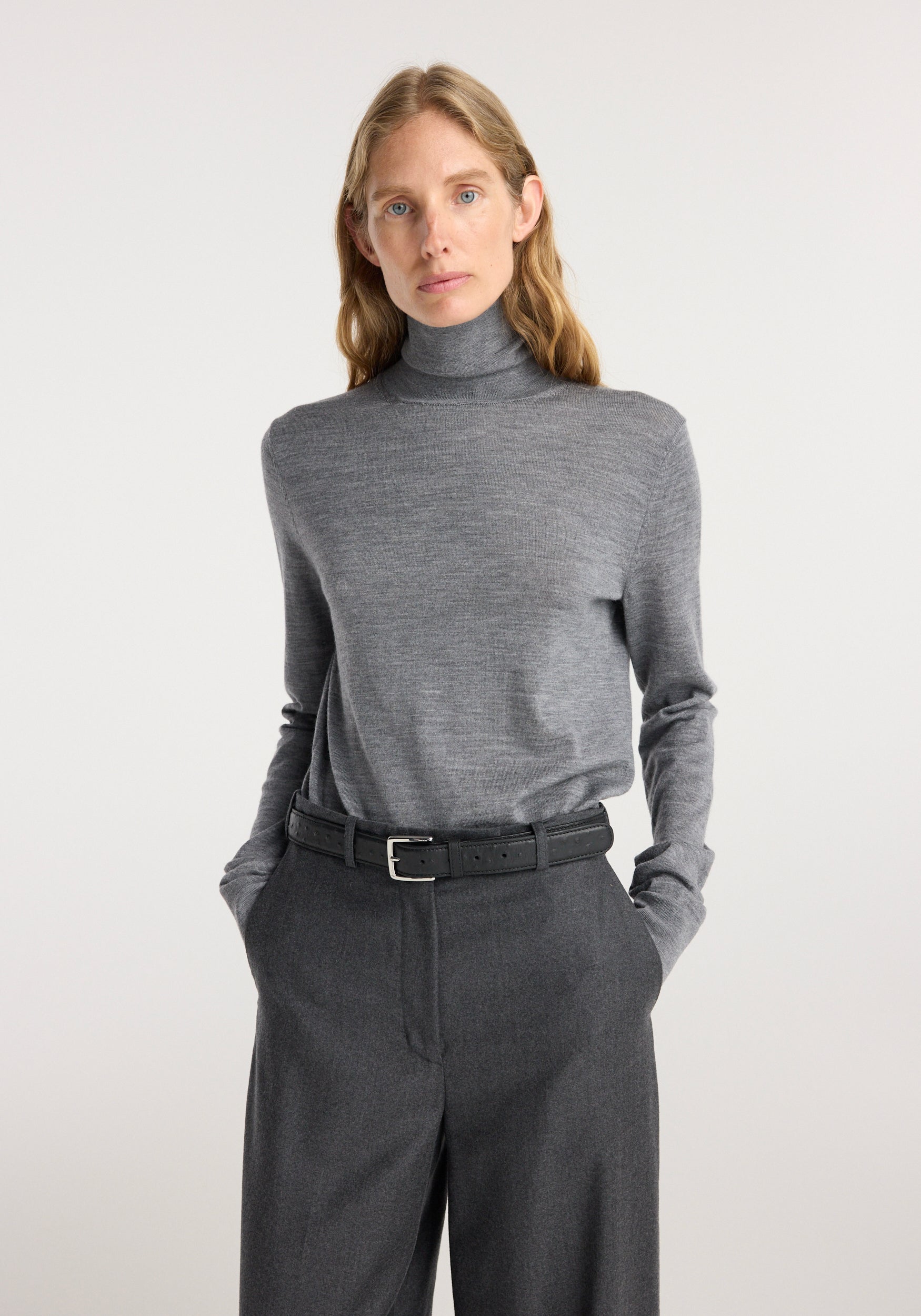 Woman wearing a mid grey melange fine knitted wool turtleneck tucked into dark grey tailored pants with a black belt against a plain light background