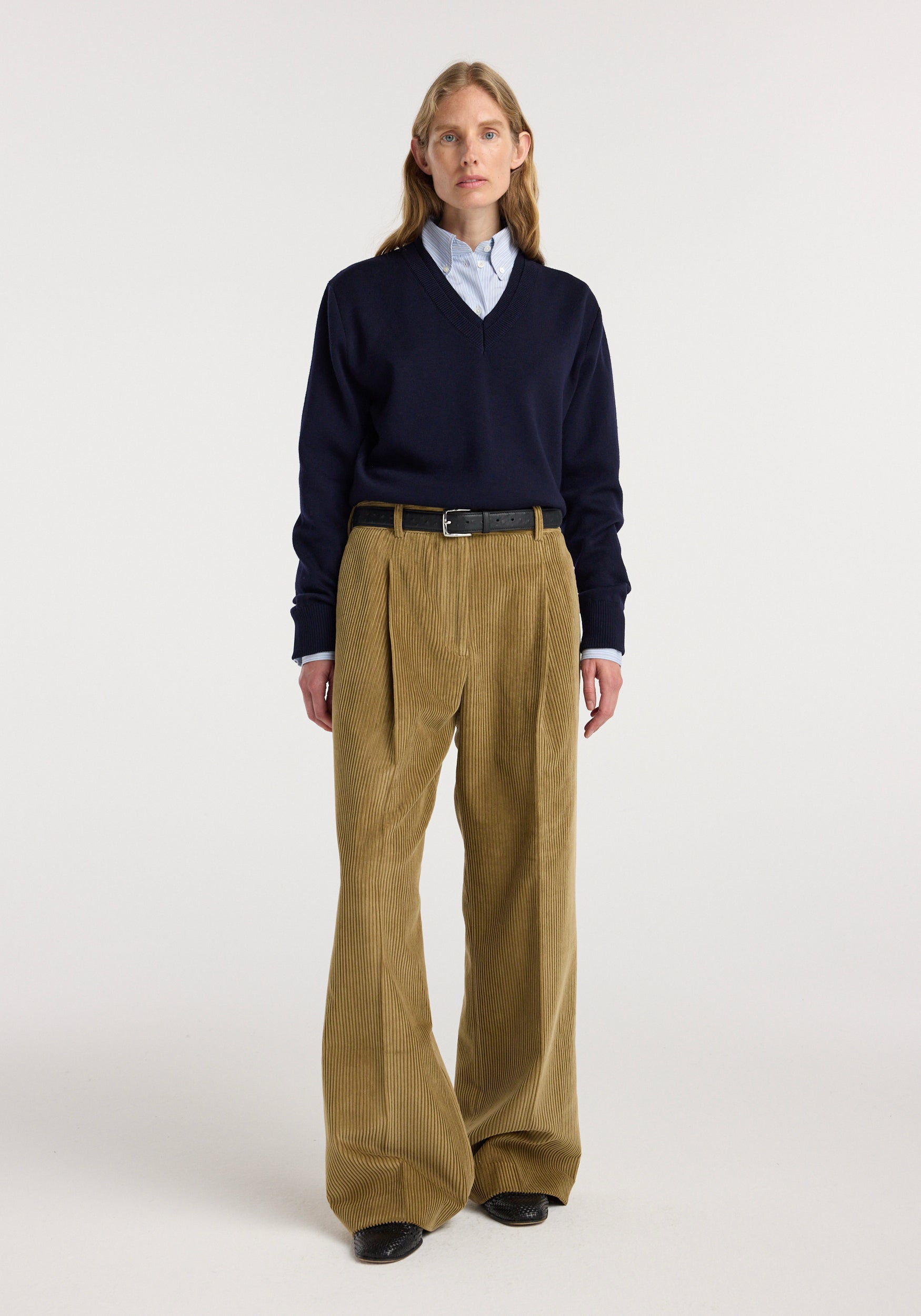 A model wearing Ted - Lower waist corduroy trousers | beige styled with a navy V-neck sweater, a light blue shirt, and black belt and shoes against a plain white background.