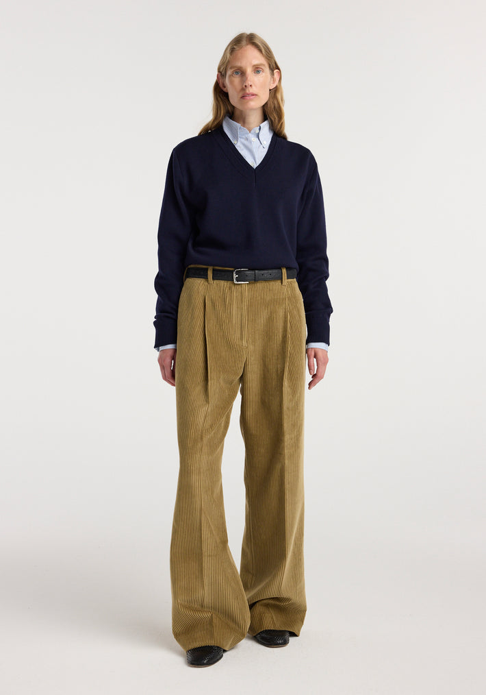 A model wearing Ted - Lower waist corduroy trousers | beige styled with a navy V-neck sweater, a light blue shirt, and black belt and shoes against a plain white background.