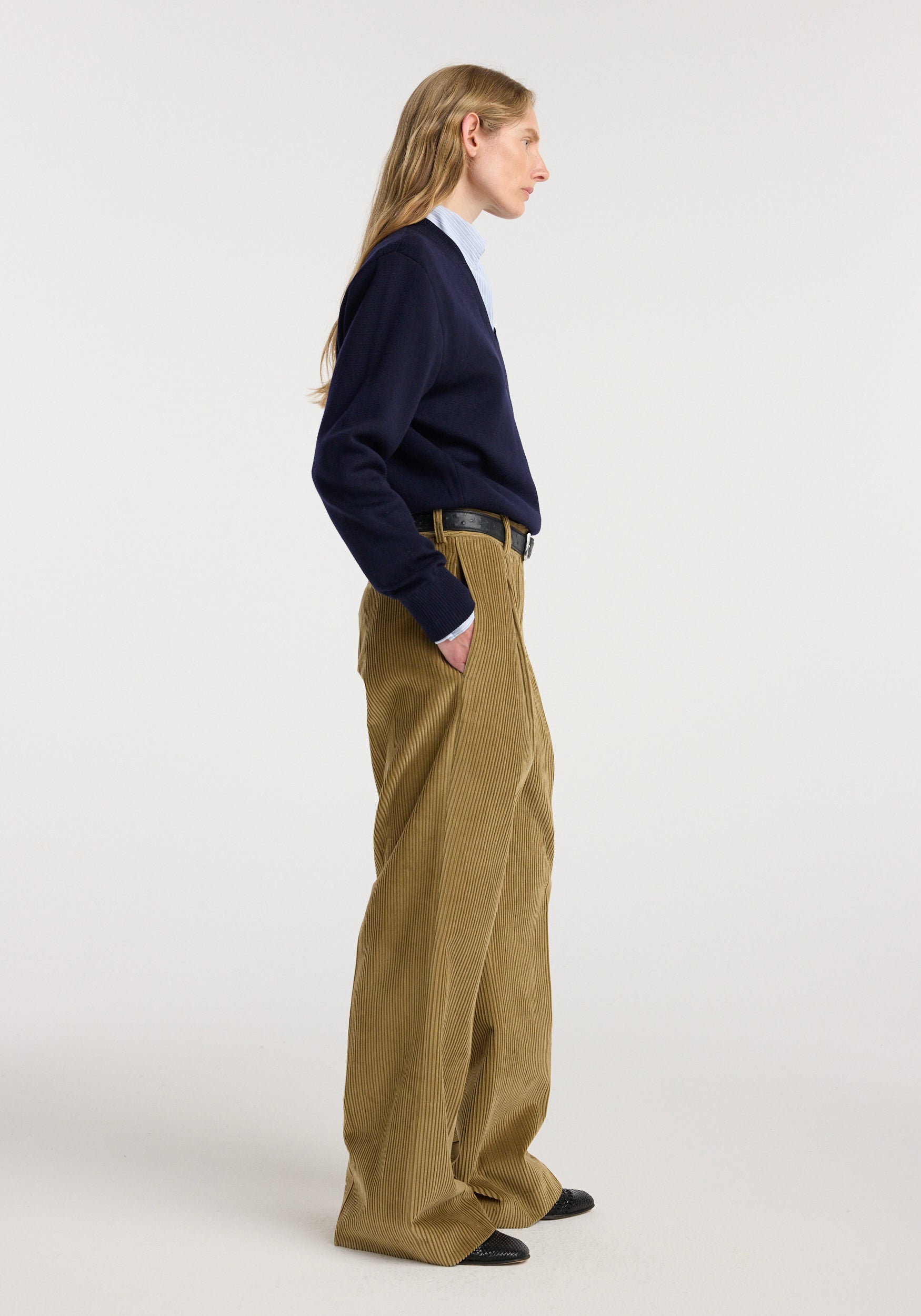Side view of a person wearing Ted - Lower waist corduroy trousers | beige with a navy sweater and light blue shirt, standing against a plain white background.