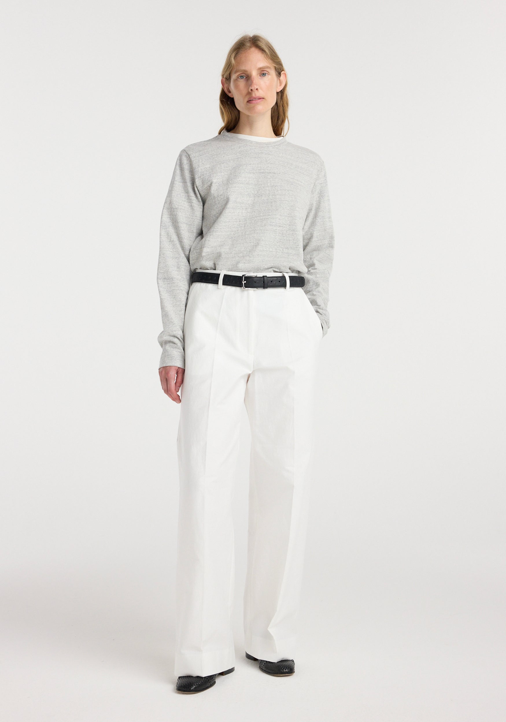 Woman wearing a grey long-sleeve sweater and classic loose fit chino | optic white pants paired with a black belt and black shoes, standing against a plain white background.
