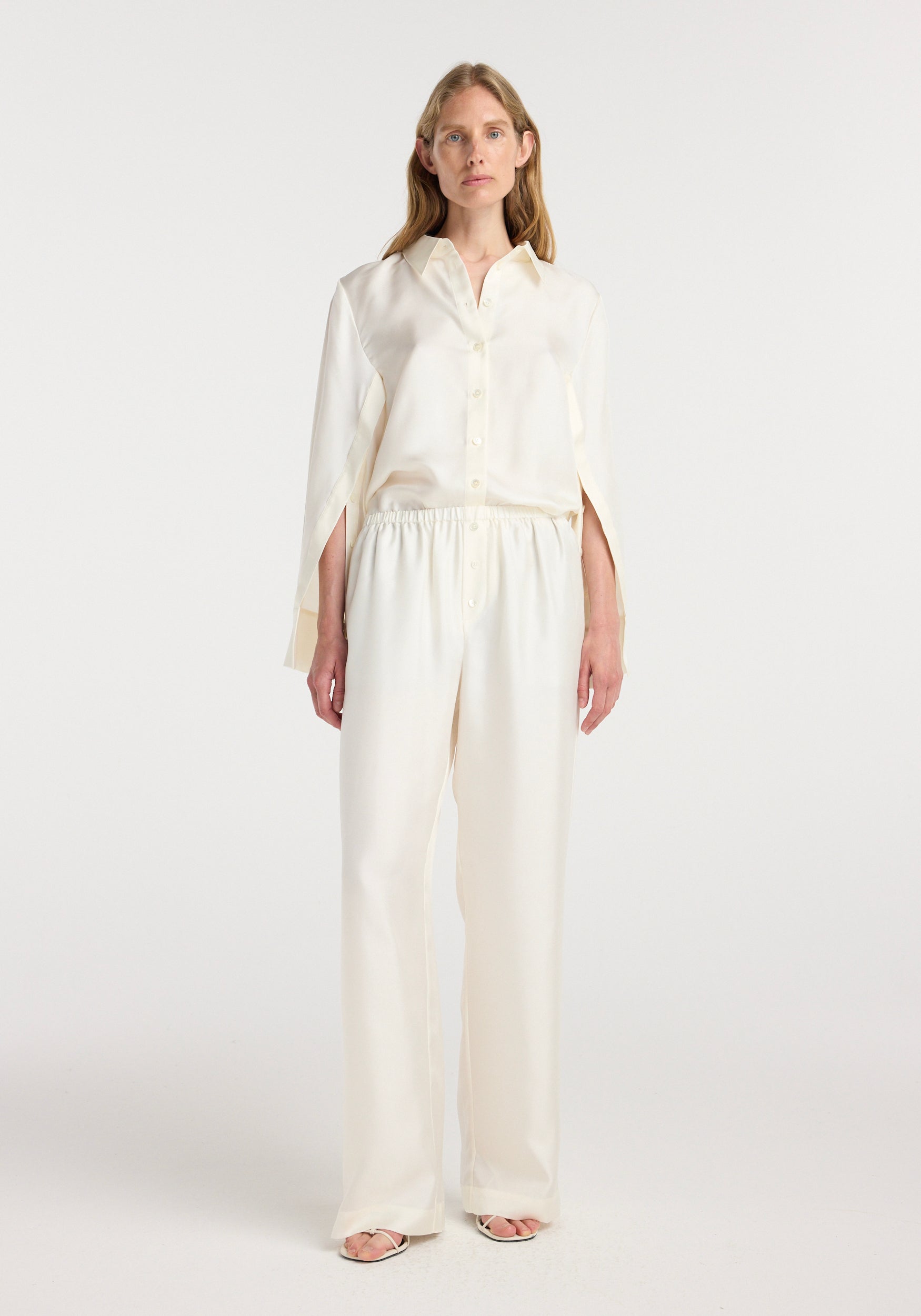 Woman wearing cream-colored silk relaxed trousers with elastic waistband, paired with a matching silk button-up shirt, standing against a plain white background.