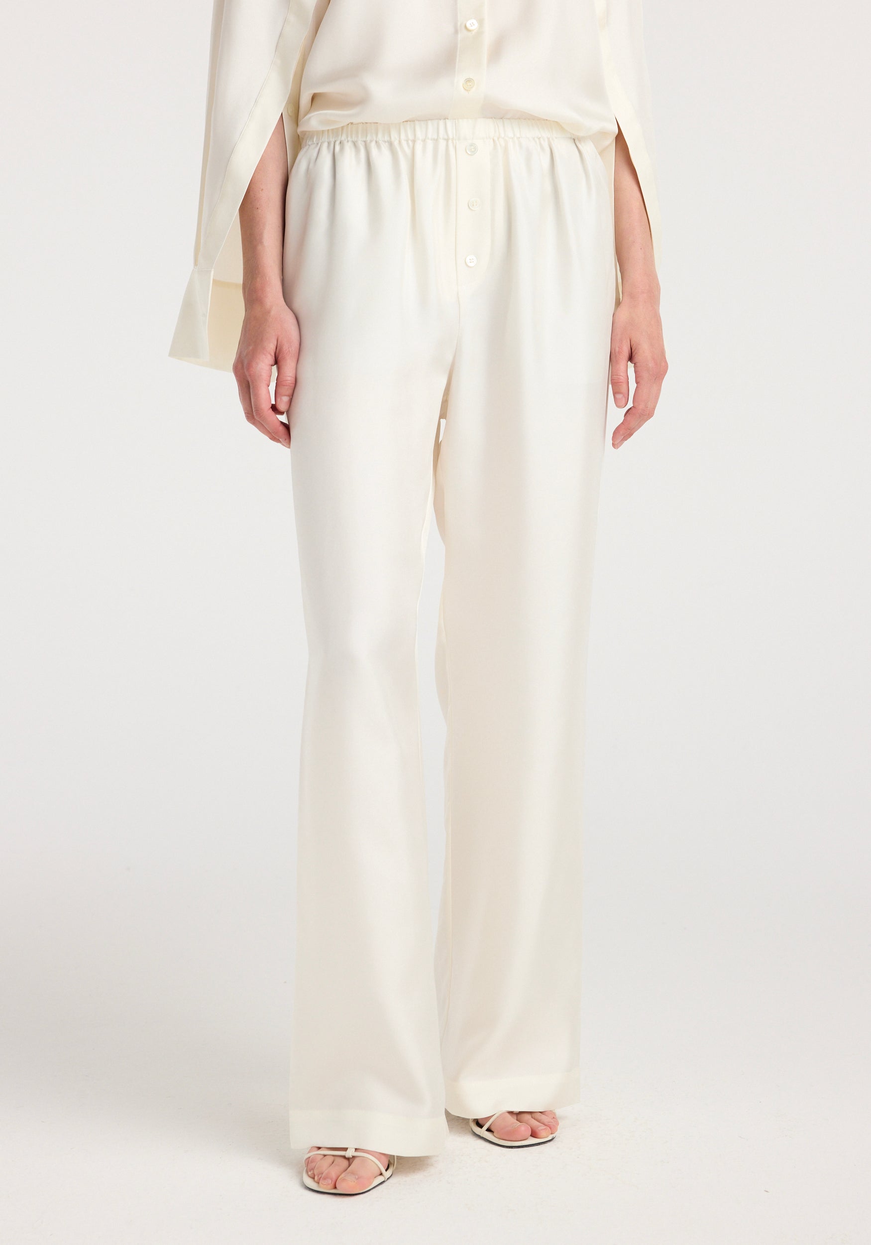 Front view of a person wearing cream-colored Silk relaxed trousers | cream with an elastic waistband, paired with a matching cream blouse and white heeled sandals.