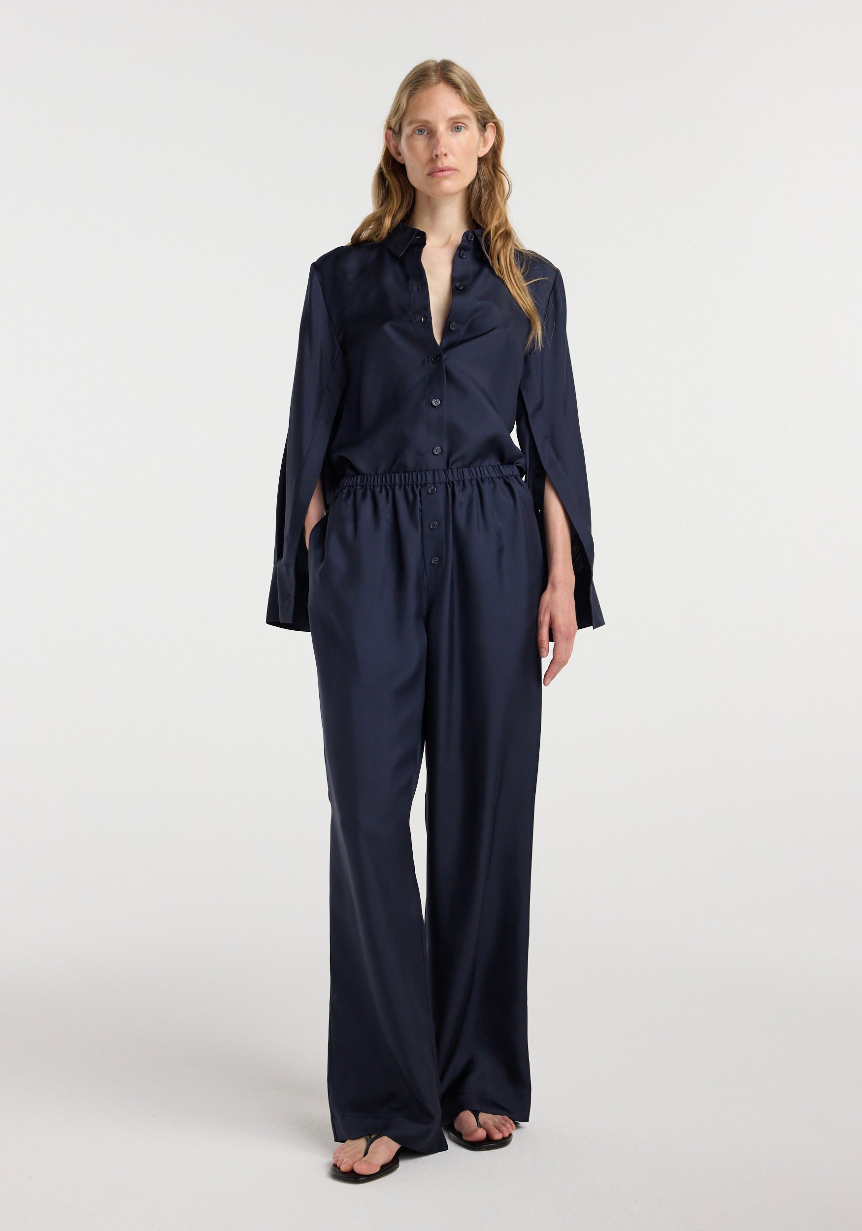 Woman wearing navy blue silk relaxed trousers | night with a matching silk button-up shirt, standing against a plain background in a minimalistic style.