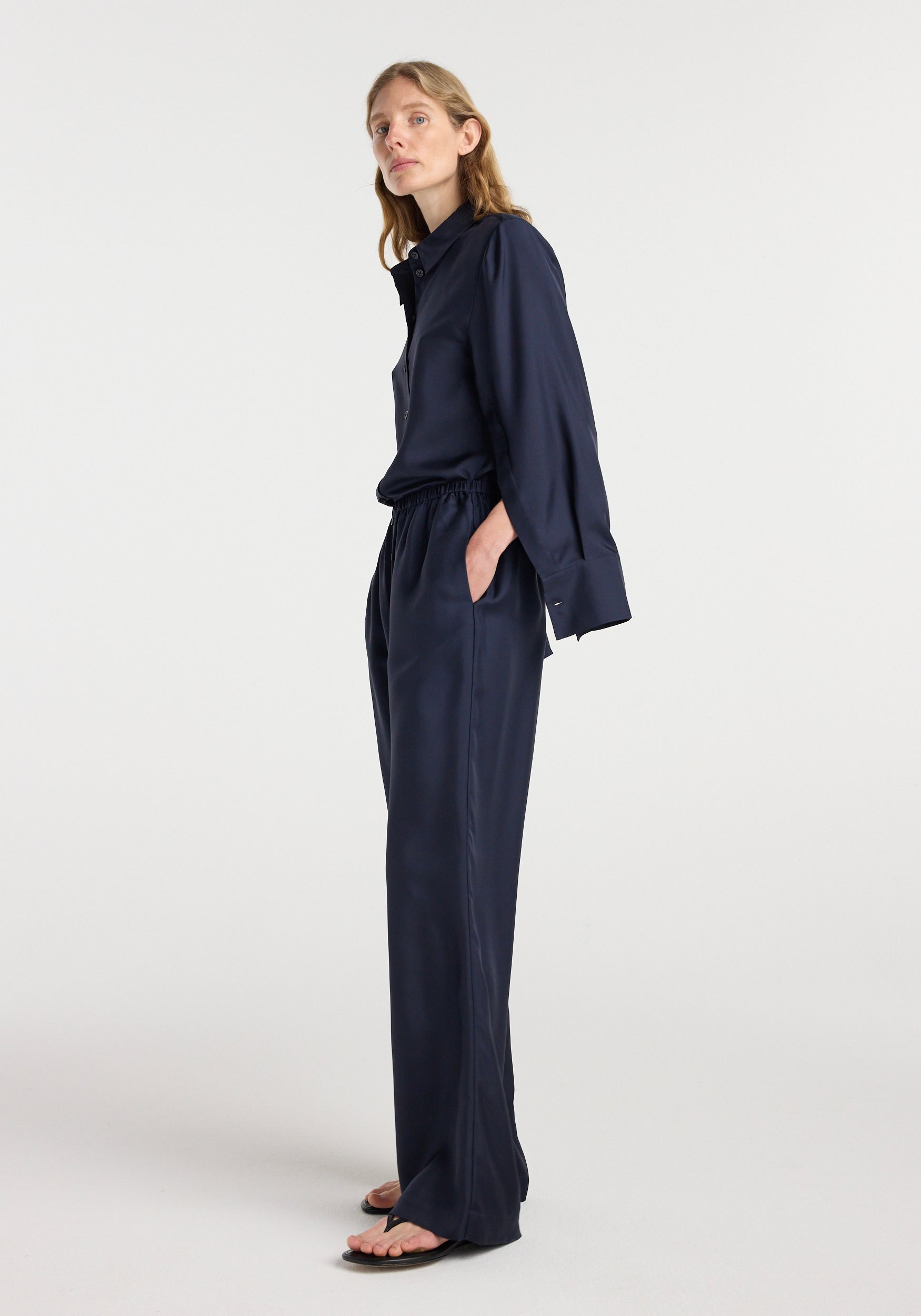 Woman wearing loose-fitting dark navy silk relaxed trousers | night paired with a matching long-sleeve silk shirt, standing in profile with hands in pockets against a plain background.