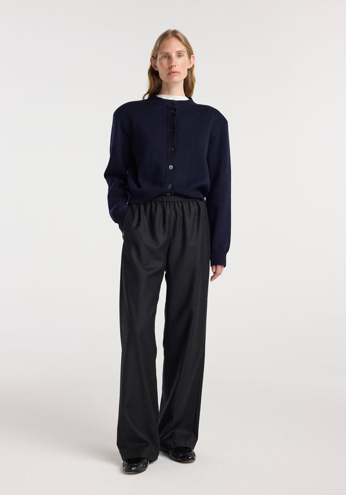 A model wearing relaxed fit trousers | anthracite melange paired with a dark navy button-up sweater and black shoes, standing against a plain white background.