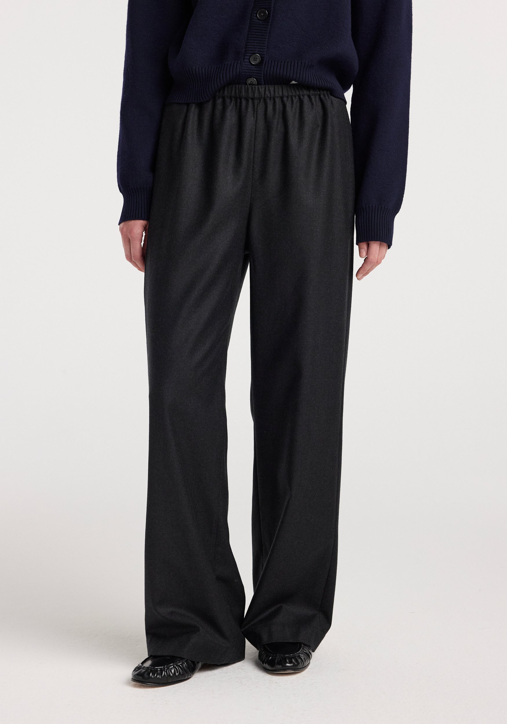 Front view of a person wearing relaxed fit trousers | anthracite melange with an elastic waistband, paired with a dark navy sweater and black flat shoes against a plain background.