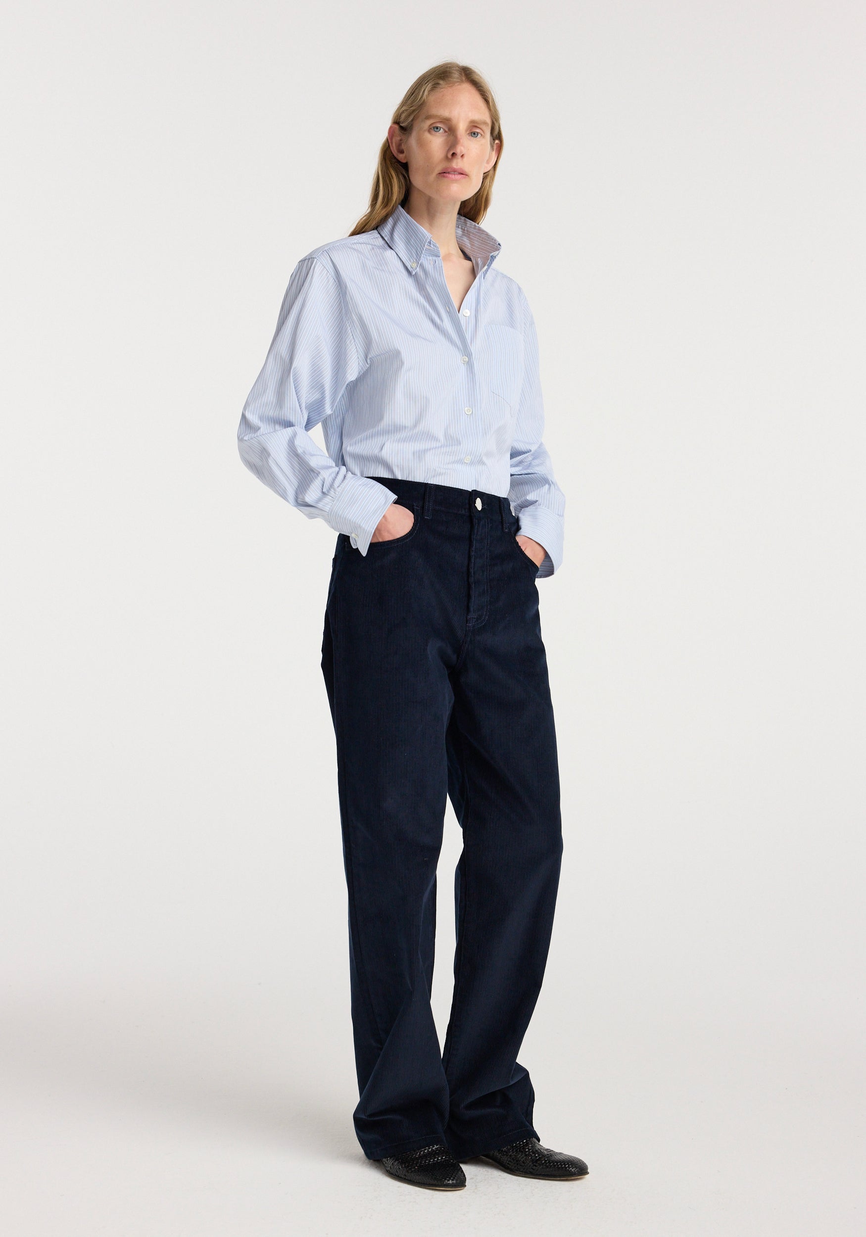 Woman wearing a light blue button-up shirt paired with Bruun - Signature baggy corduroy | night in dark navy, standing with hands in pockets against a plain white background.