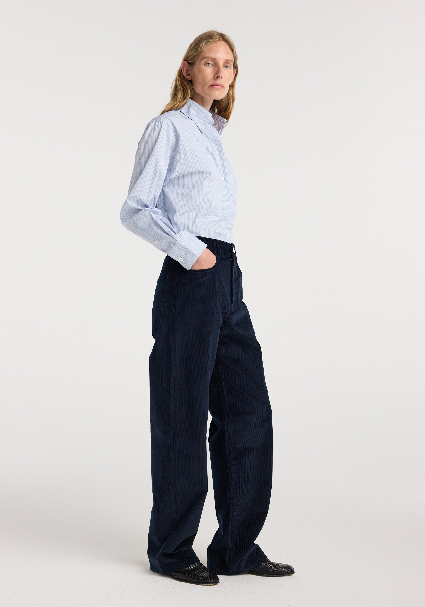Female model wearing Bruun - Signature baggy corduroy | night colored loose-fitting pants paired with a light blue button-up shirt and black shoes against a plain white background.