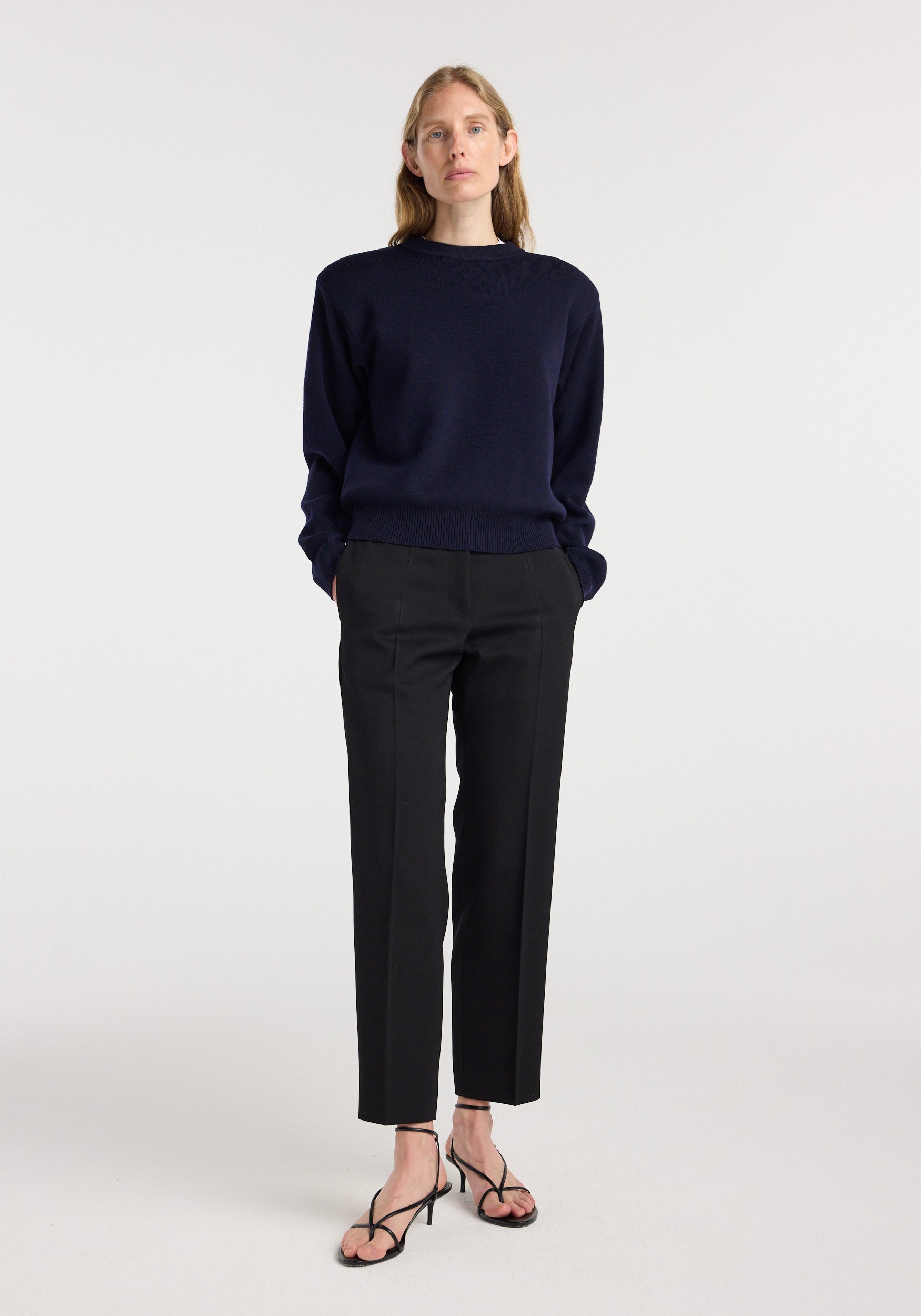 Woman wearing Mies - Cigarette leg tailored trousers | black paired with a dark navy sweater and black strappy heeled sandals, standing against a plain light background.