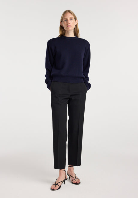 Woman wearing Mies - Cigarette leg tailored trousers | black paired with a dark navy sweater and black strappy heeled sandals, standing against a plain light background.