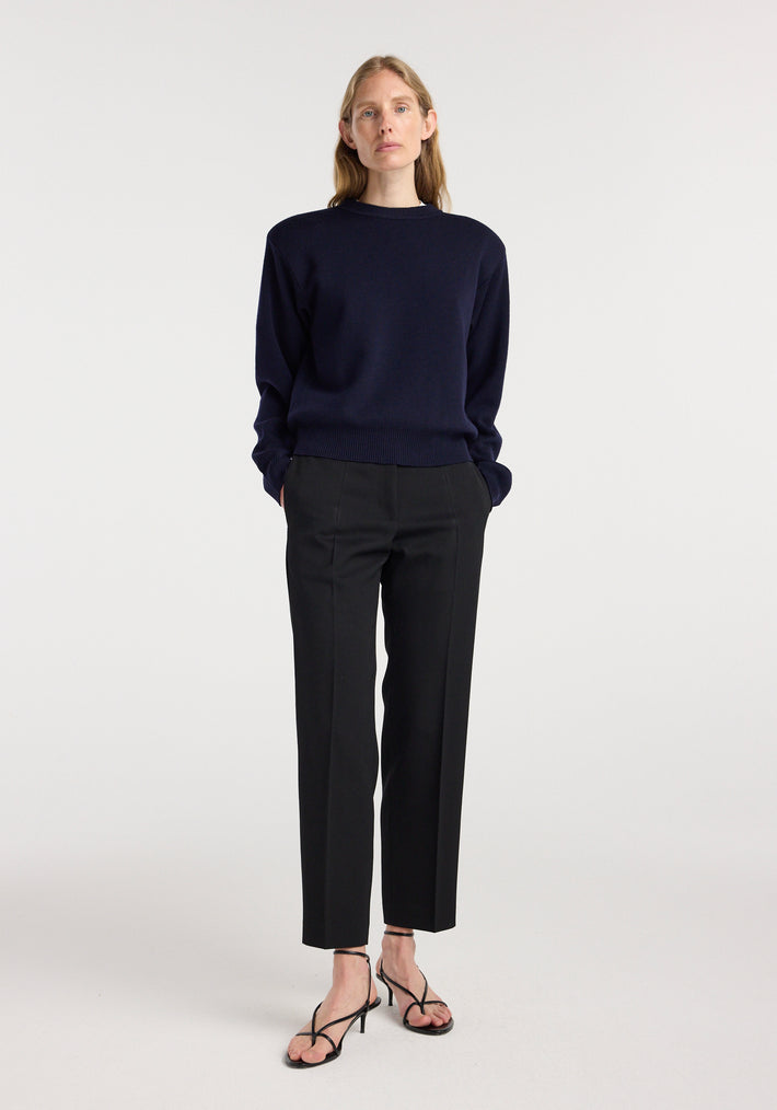 Woman wearing Mies - Cigarette leg tailored trousers | black paired with a dark navy sweater and black strappy heeled sandals, standing against a plain light background.
