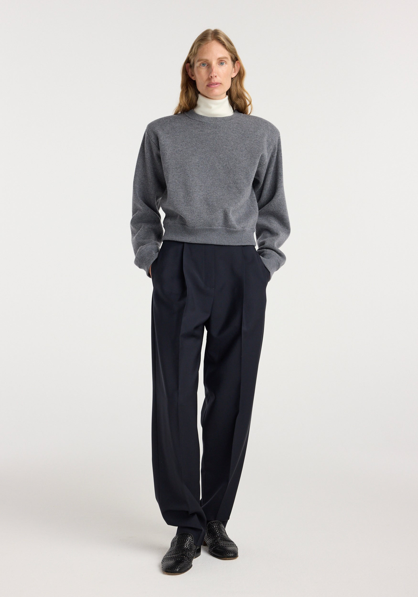 Woman wearing Daria - Single pleat tapered trousers | navy paired with a grey sweater and white turtleneck, standing against a plain white background.