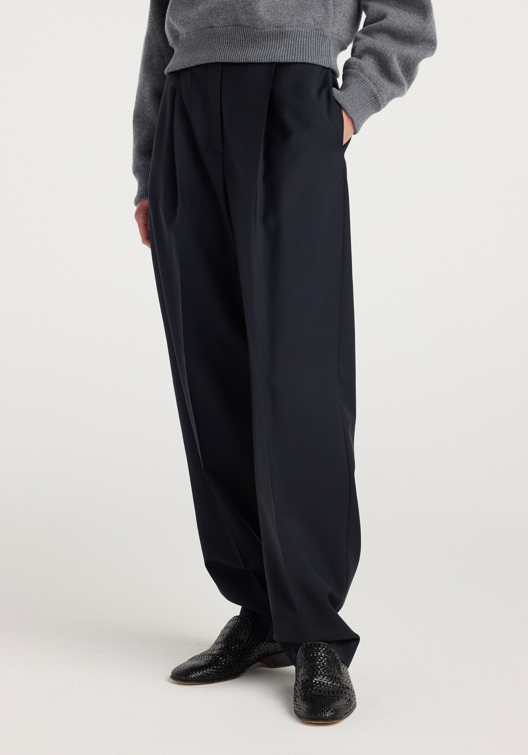 Close-up of a person wearing Daria - Single pleat tapered trousers | navy, styled with a gray sweater and black textured shoes against a plain background.
