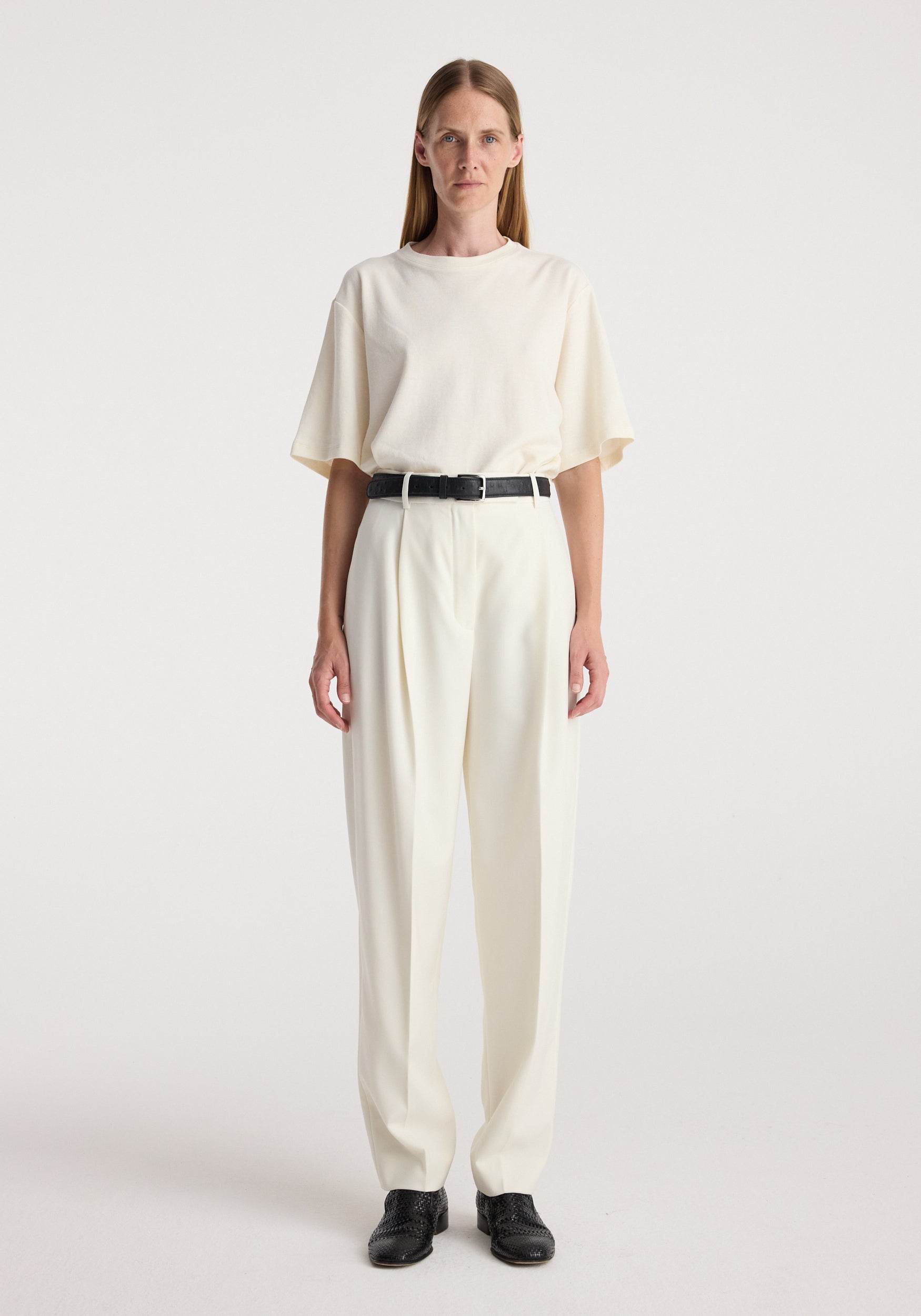 Woman wearing Daria - Single pleat tapered trousers | off white paired with a loose off-white top and black belt, standing against a plain white background.