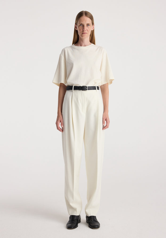 Woman wearing Daria - Single pleat tapered trousers | off white paired with a loose off-white top and black belt, standing against a plain white background.