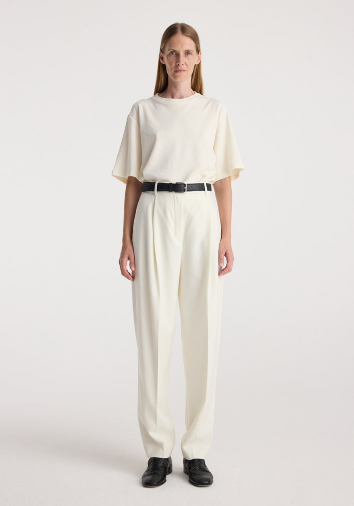 Woman wearing Daria - Single pleat tapered trousers | off white paired with a loose off-white top and black belt, standing against a plain white background.