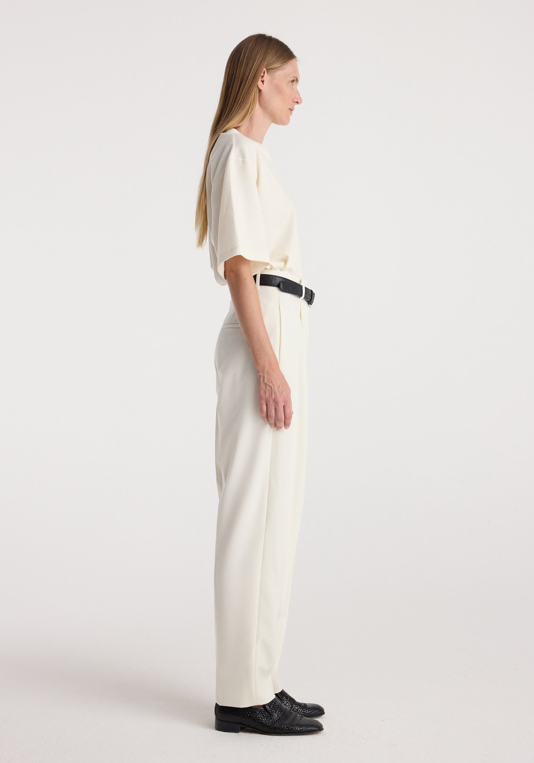 Profile view of a woman wearing Daria - Single pleat tapered trousers | off white paired with a matching short sleeve top, black belt, and black shoes against a plain background.