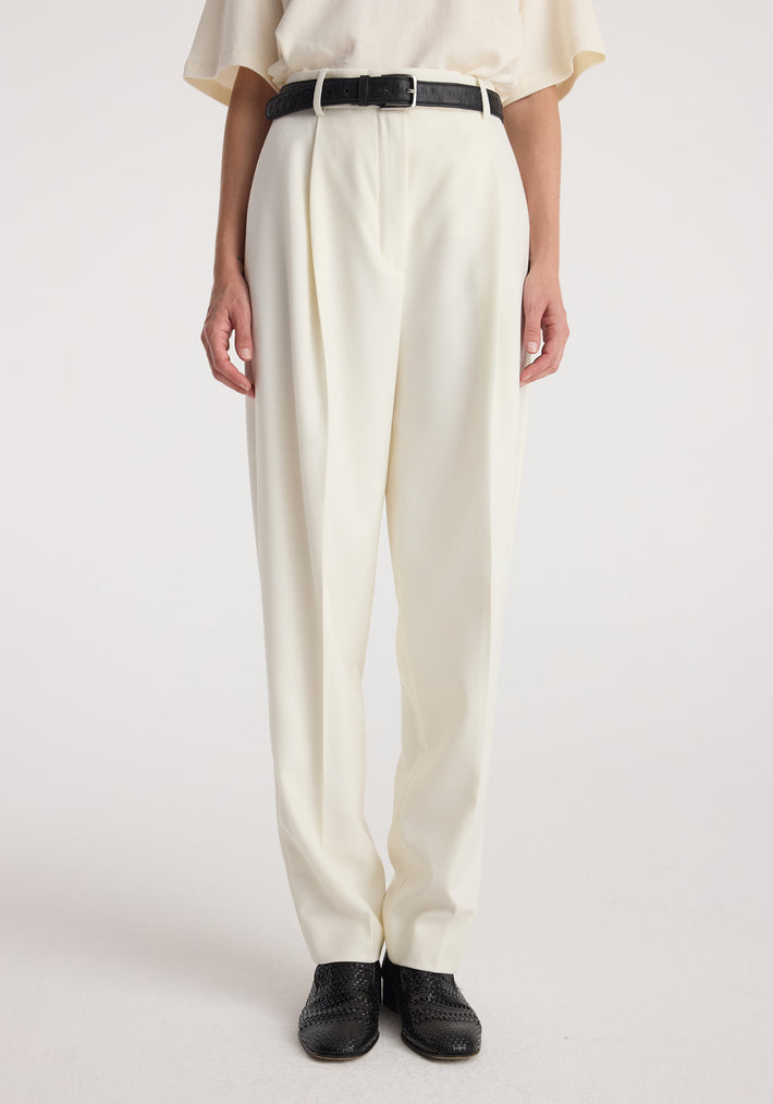 Front view of a person wearing Daria - Single pleat tapered trousers | off white paired with a black belt and black shoes against a plain background.