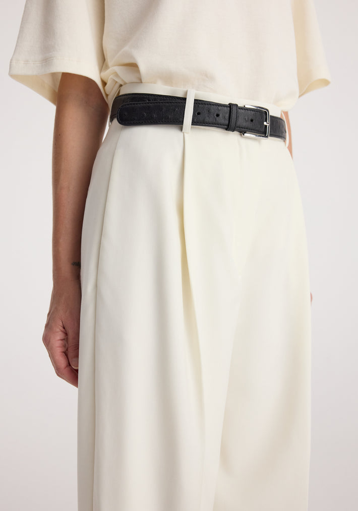 Close-up of a person wearing Daria - Single pleat tapered trousers | off white with a black belt and a loose off-white top, showcasing the trousers' pleated design and tapered fit.