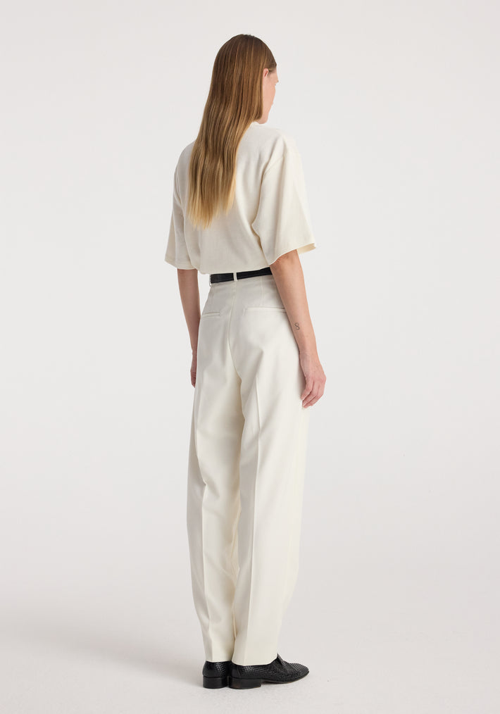 Woman wearing off white Daria - Single pleat tapered trousers standing with back facing the camera, paired with a loose short-sleeve top and black shoes against a plain background.