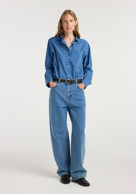 Woman wearing Bruun - Signature baggy denim | heritage blue paired with a blue button-up shirt, black belt, and black shoes, standing against a white background.