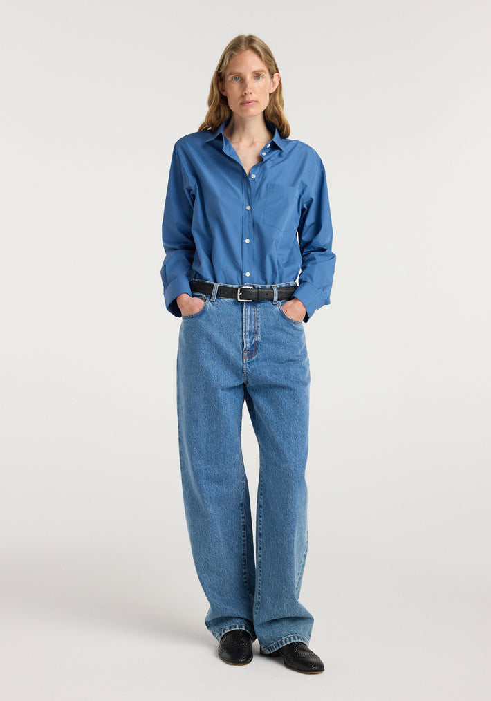 Woman wearing Bruun - Signature baggy denim | heritage blue paired with a blue button-up shirt, black belt, and black shoes, standing against a white background.