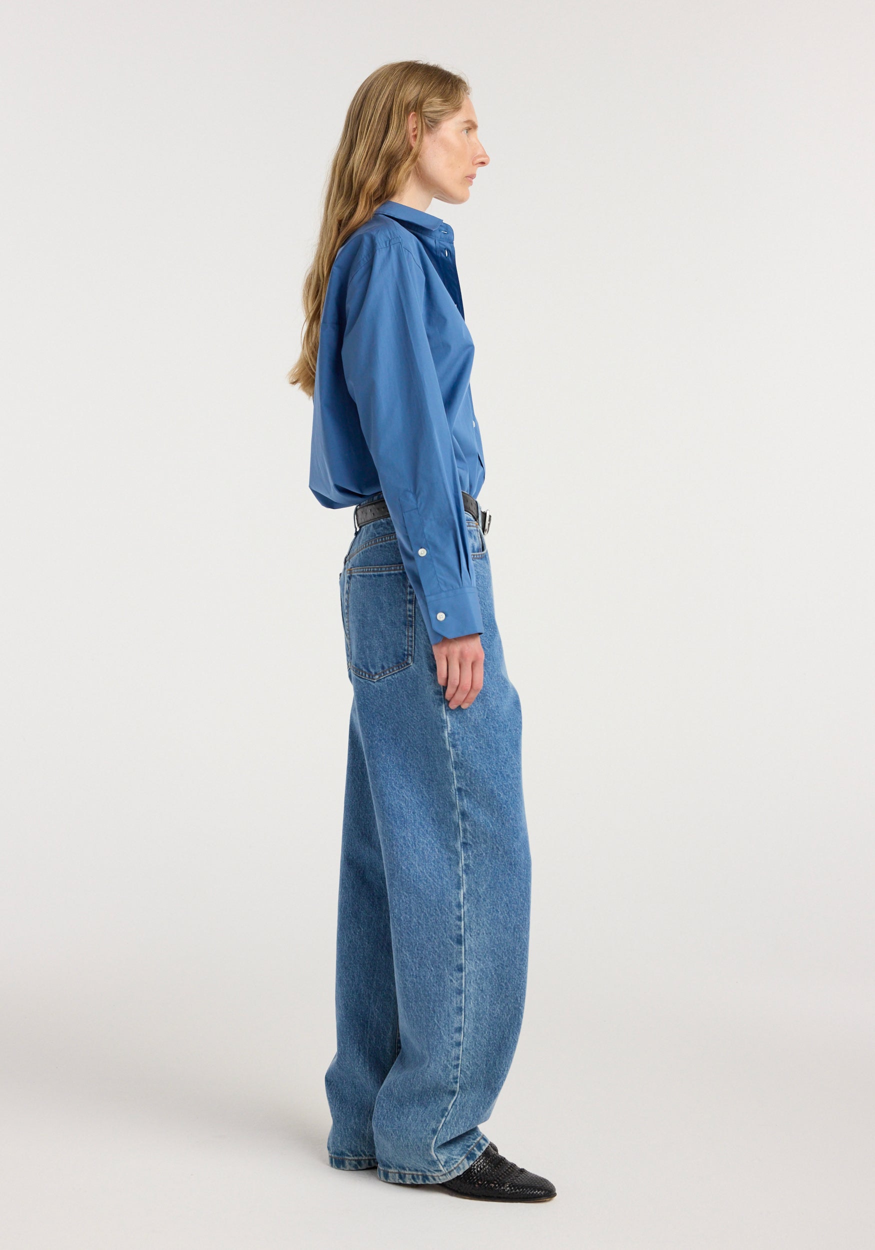 Side profile of a woman wearing Bruun - Signature baggy denim | heritage blue paired with a blue button-up shirt and black shoes against a plain background.