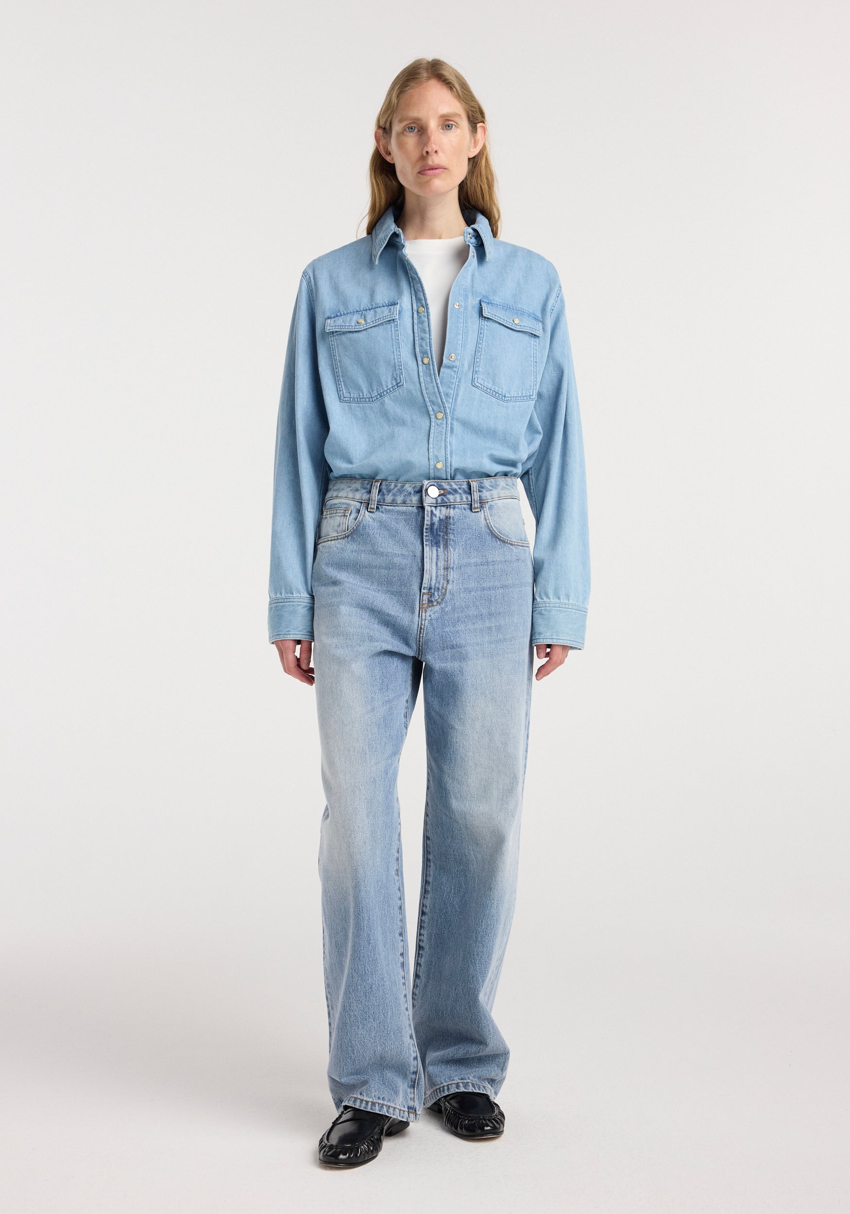 Woman wearing classic blue Bruun - Signature baggy denim with a light blue denim shirt and black shoes, standing against a plain white background.