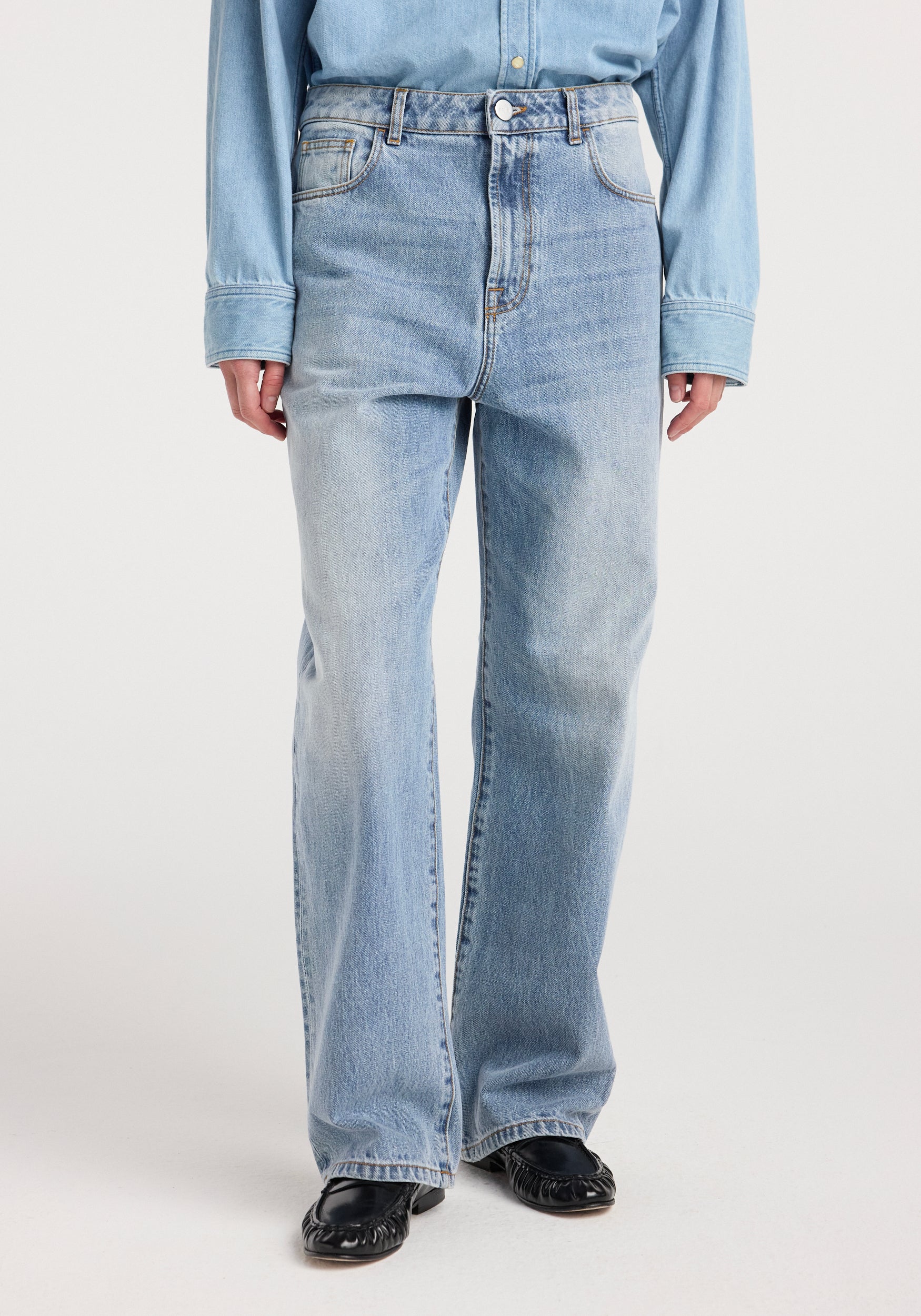Front view of a person wearing Bruun - Signature baggy denim | classic blue with a relaxed fit, paired with a light blue button-up shirt and black shoes on a white background.